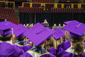 Auditorium of graduates in purple robes and caps listening to speak on stage